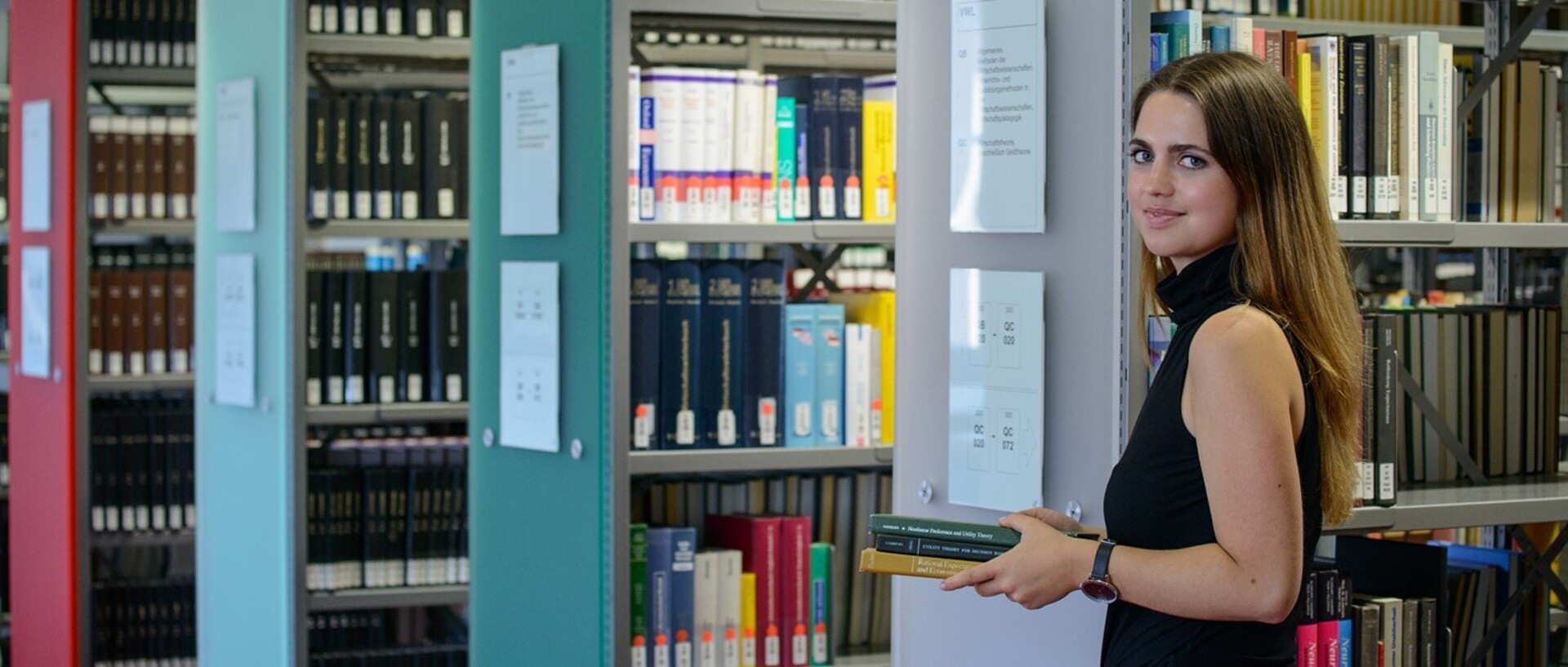 A student stands in front of shelves in the “Ehrenhof”-Library. She is holding a pile of books in her hand.