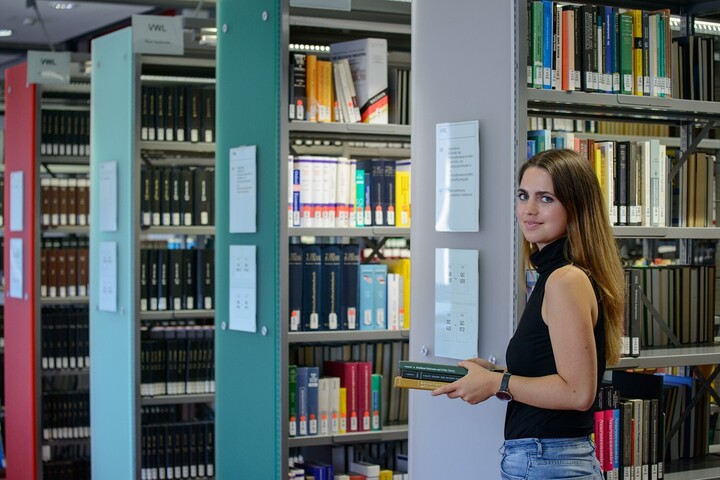 A student stands in front of shelves in the “Ehrenhof”-Library. She is holding a pile of books in her hand.