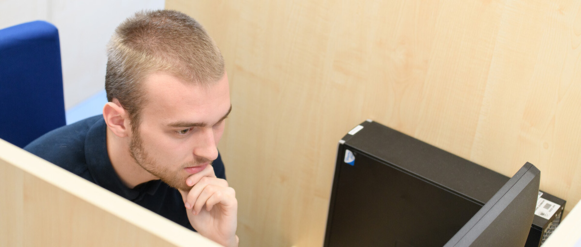Ein junger Mann sitzt nachdenklich in einer abgetrennten Arbeitskabine vor einem Computerbildschirm. Seine Hand stützt sein Kinn, während er sich konzentriert auf die angezeigten Inhalte fokussiert.