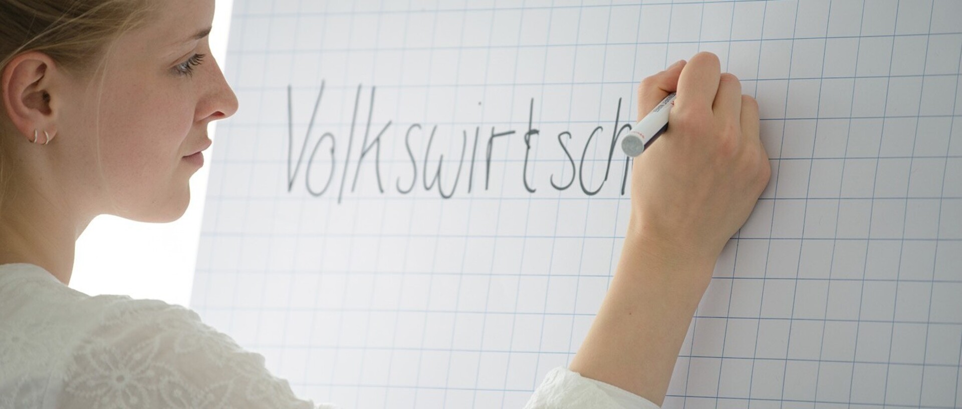 Eine Frau schreibt mit einem schwarzen Filzstift auf ein Flipchart mit kariertem Papier das Wort „Volkswirtschaftslehre“. Sie hat blonde Haare, die zu einem Zopf gebunden sind, trägt zwei schmale goldene Kreolen und eine weiße T-Shirtbluse.