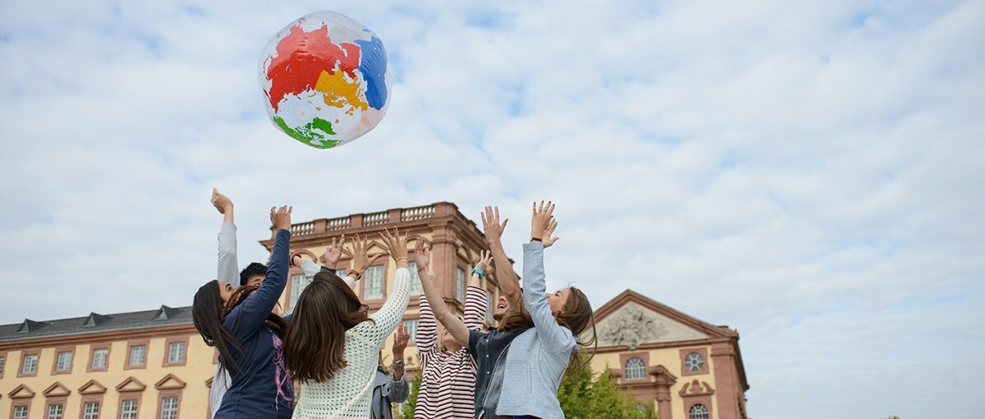 Eine Gruppe Studierender wirft eine Weltkugel in die Luft. Im Hintergrund befindet sich die Universität Mannheim.