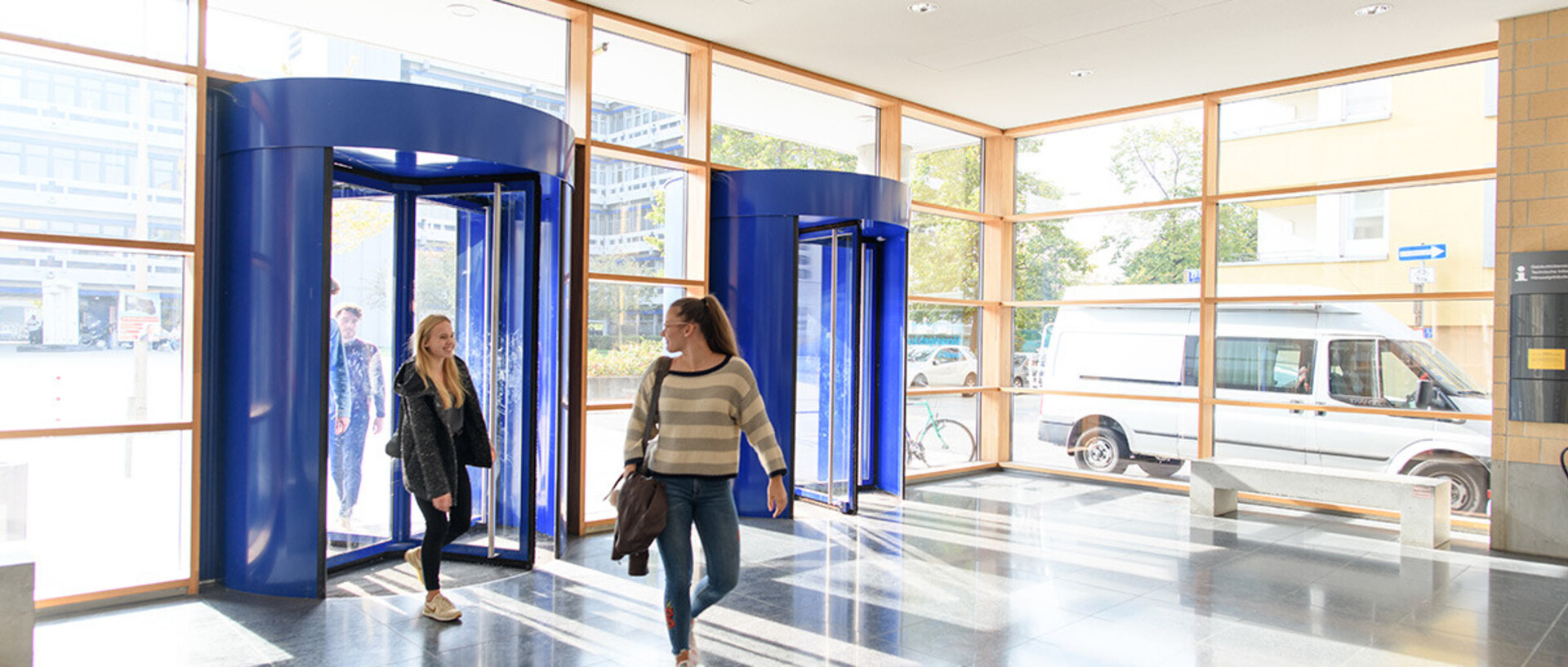 Zwei Studentinnen betreten das Foyer von Gebäude B6 durch die dunkelblau eingefassten Drehtüren.