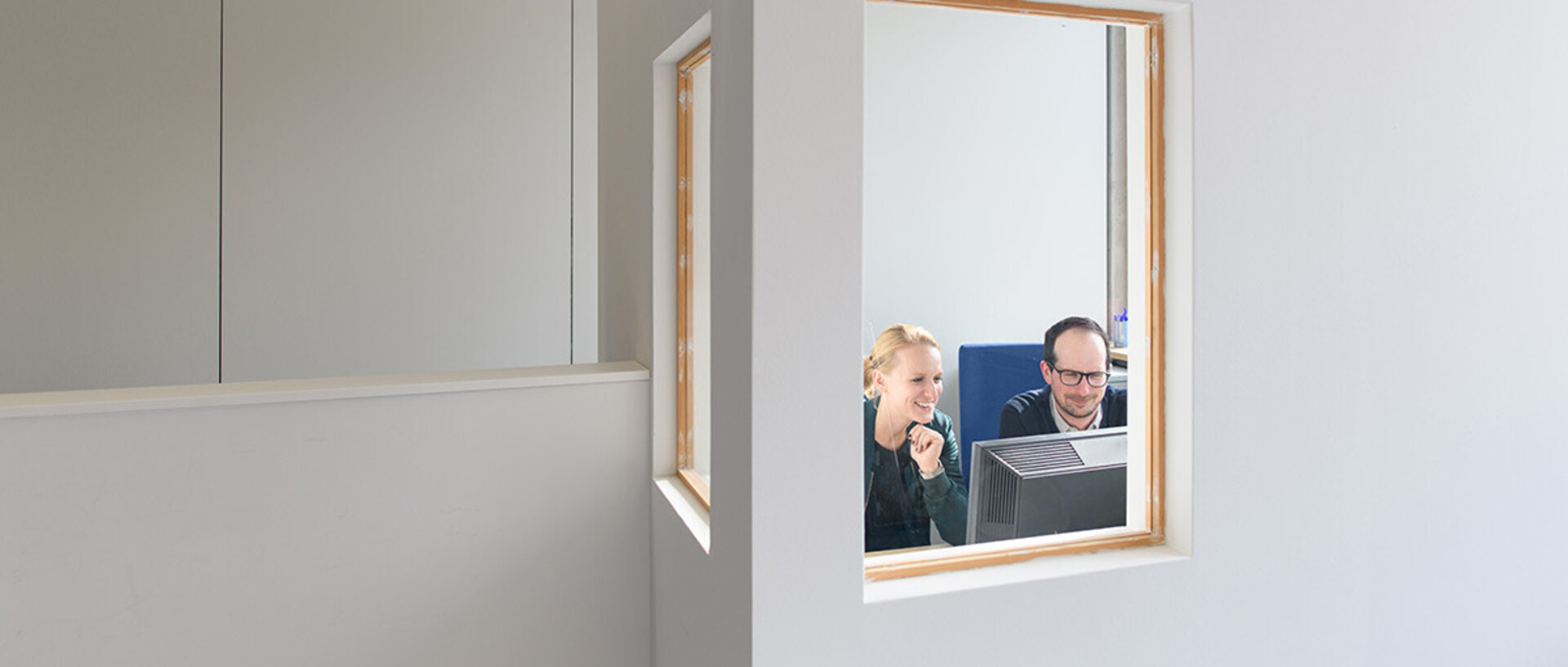 Eine Frau und ein Mann sind durch ein Fenster in einem separaten Raum zu sehen, während sie gemeinsam auf einen Computerbildschirm schauen.