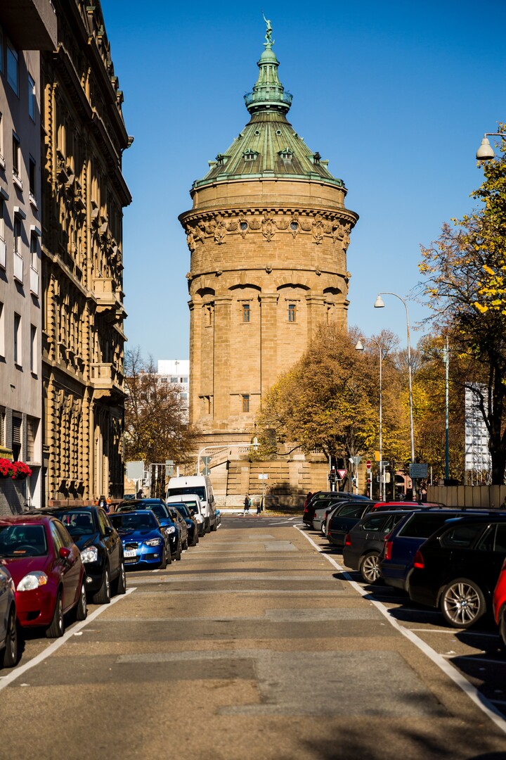 Blick auf den Mannheimer Wasserturm. Am linken und rechten Bildrand parken viele Autos und am linken Rand erkennt man eine Häuserfront. Rechts stehen einige Bäume und Straßenlaternen.