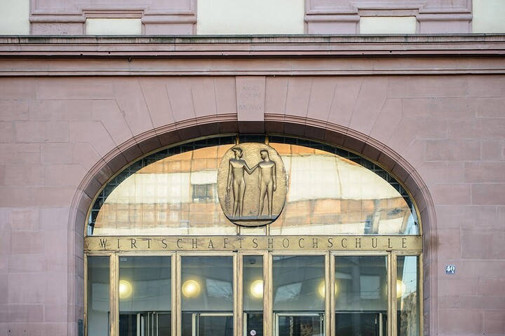 Hauptportal der Universität Mannheim. Über dem Eingang steht „Wirtschaftshochschule“, darüber befindet sich ein ovalförmiges, goldenes Emblem, das zwei Menschen abbildet.