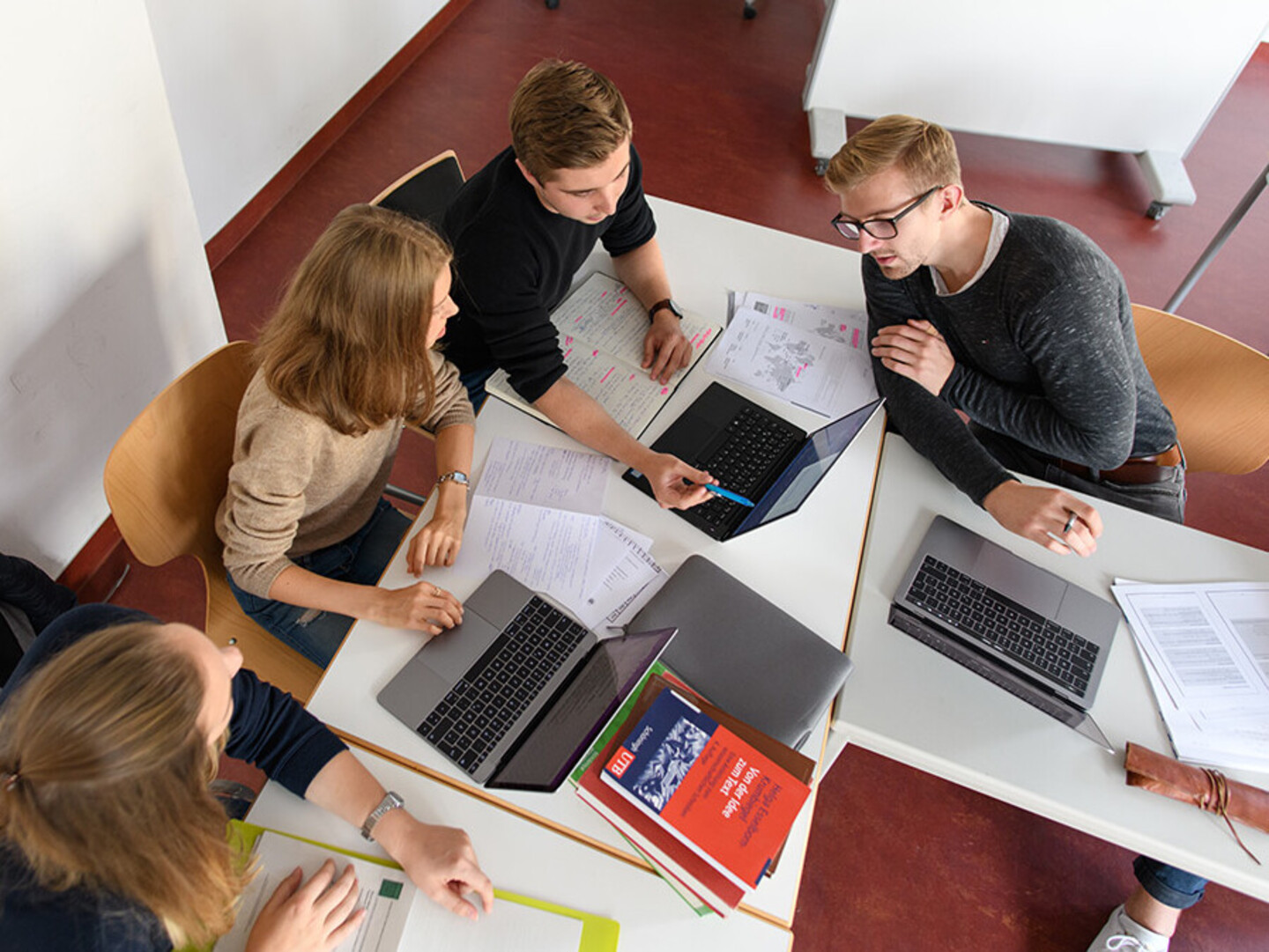 Eine Gruppe von vier Personen sitzt um einen Tisch und arbeitet gemeinsam an einem Projekt. Auf dem Tisch liegen Laptops, Notizbücher und Dokumente.