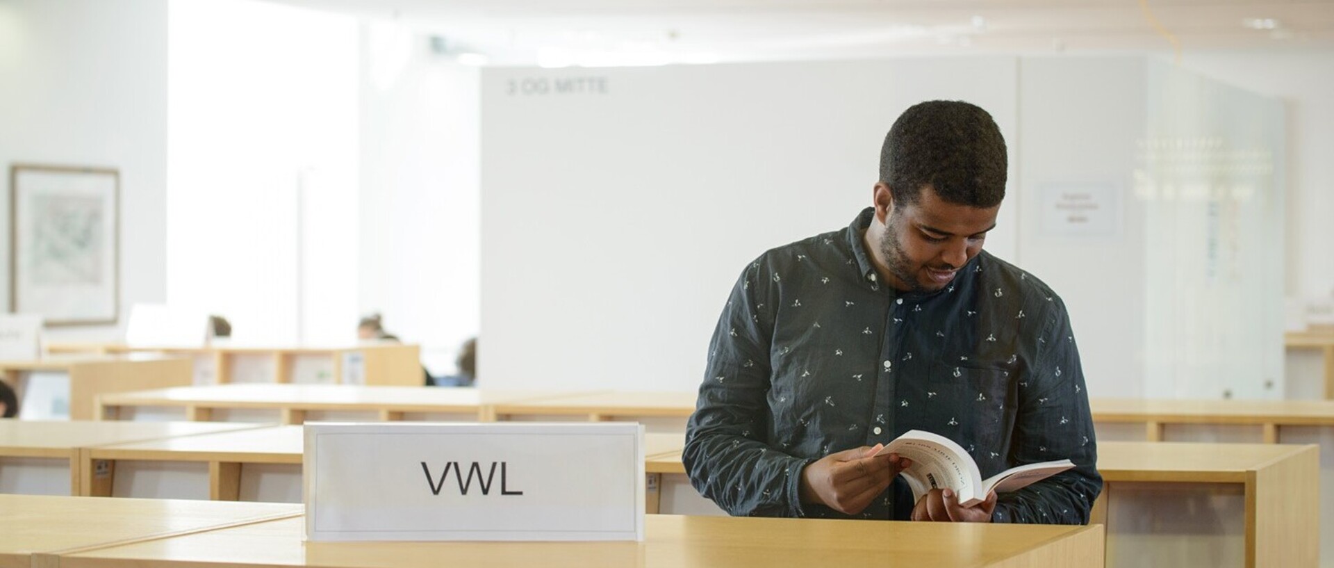 A student with short black hair and a dark-colored shirt with white dots is holding an open book in his hands. In front of him, on a low shelf unit, is a sign reading ‘VWL’ [Economics].