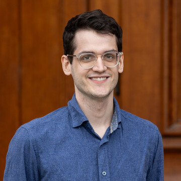 He is wearing a dark blue, jeans-like shirt and is standing in front of an entrance to Mannheim Palace.