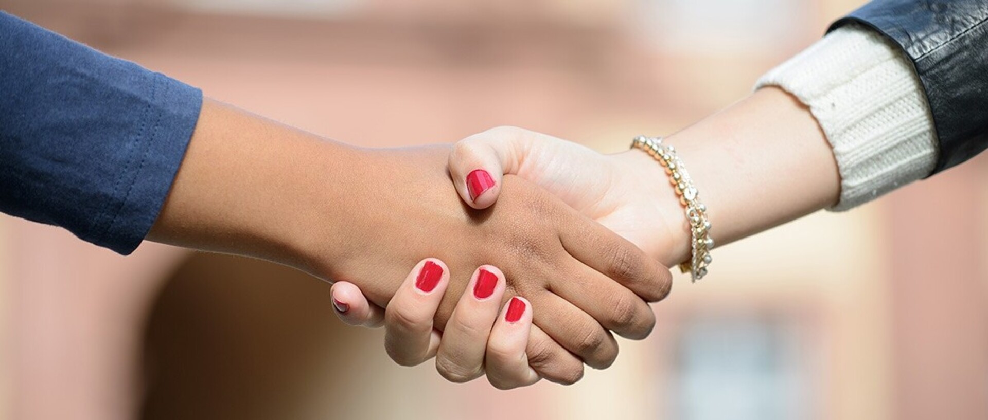 Zwei Hände werden gereicht. Die rechte Hand hat rot lackierte Fingernägel und ein goldenes Armband wird um das Handgelenk getragen.