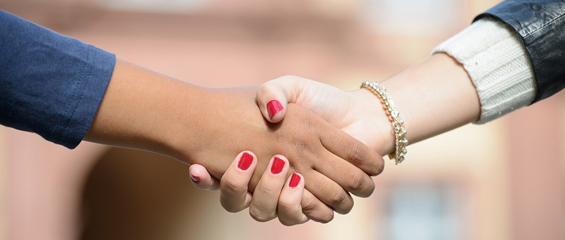 Zwei Hände werden gereicht. Die rechte Hand hat rot lackierte Fingernägel und ein goldenes Armband wird um das Handgelenk getragen.
