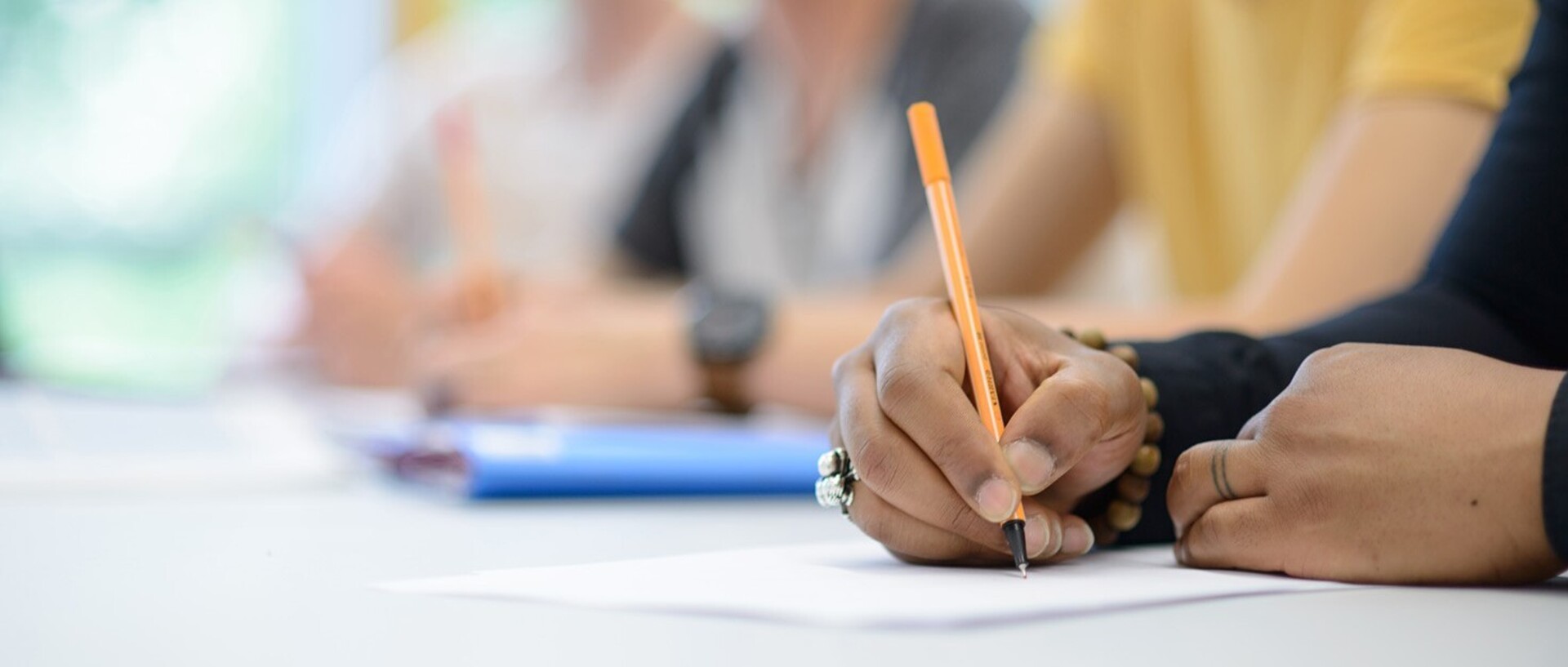 A person is writing on a white sheet of paper. She is holding an orange pen in her hand.