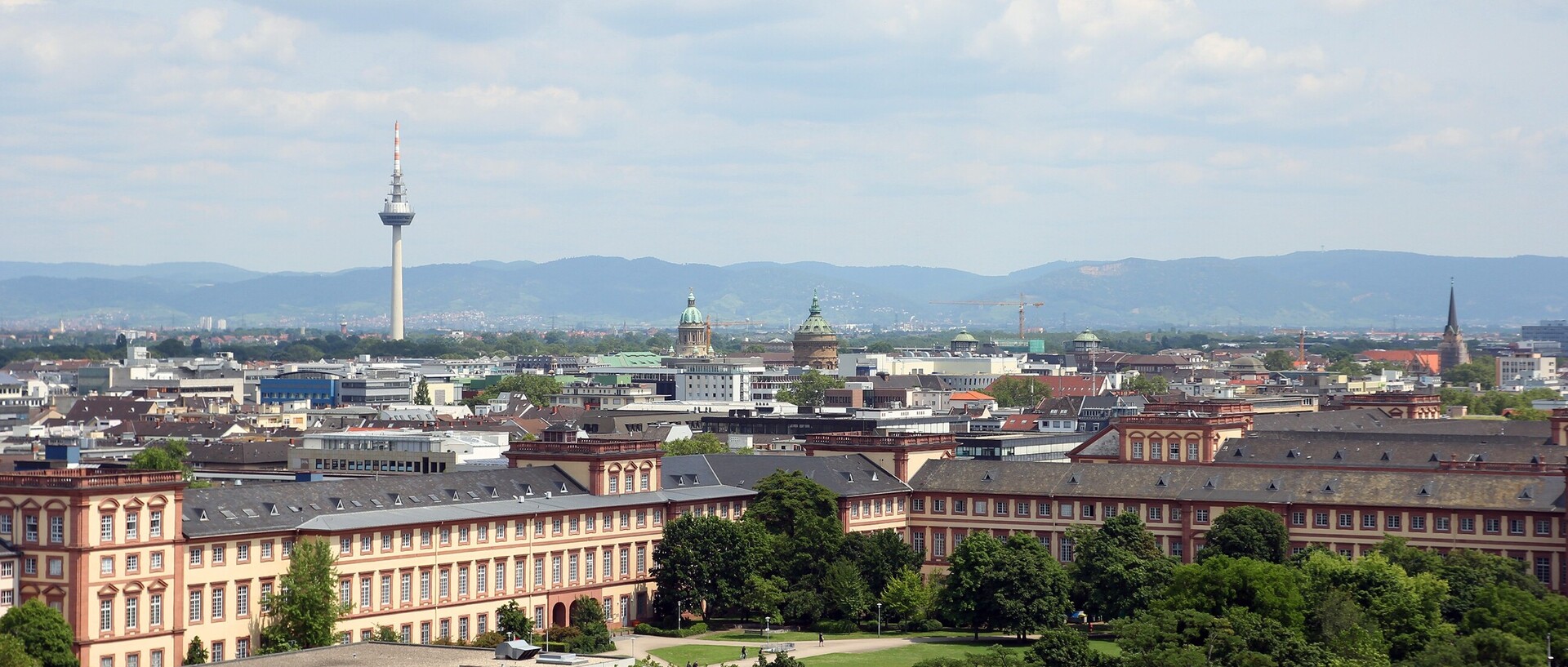 Blick über die Stadt Mannheim. Im Vordergrund ist das Schloss und die Mensawiese zu erkennen. Im Hintergrund erstrecken sich die Stadt und dahinter einige Hügel. Markant befindet sich hinten links im Bild der Fernsehturm der Stadt Mannheim.