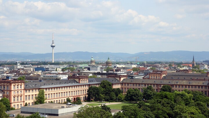 Blick über die Stadt Mannheim. Im Vordergrund ist das Schloss und die Mensawiese zu erkennen. Im Hintergrund erstrecken sich die Stadt und dahinter einige Hügel. Markant befindet sich hinten links im Bild der Fernsehturm der Stadt Mannheim.