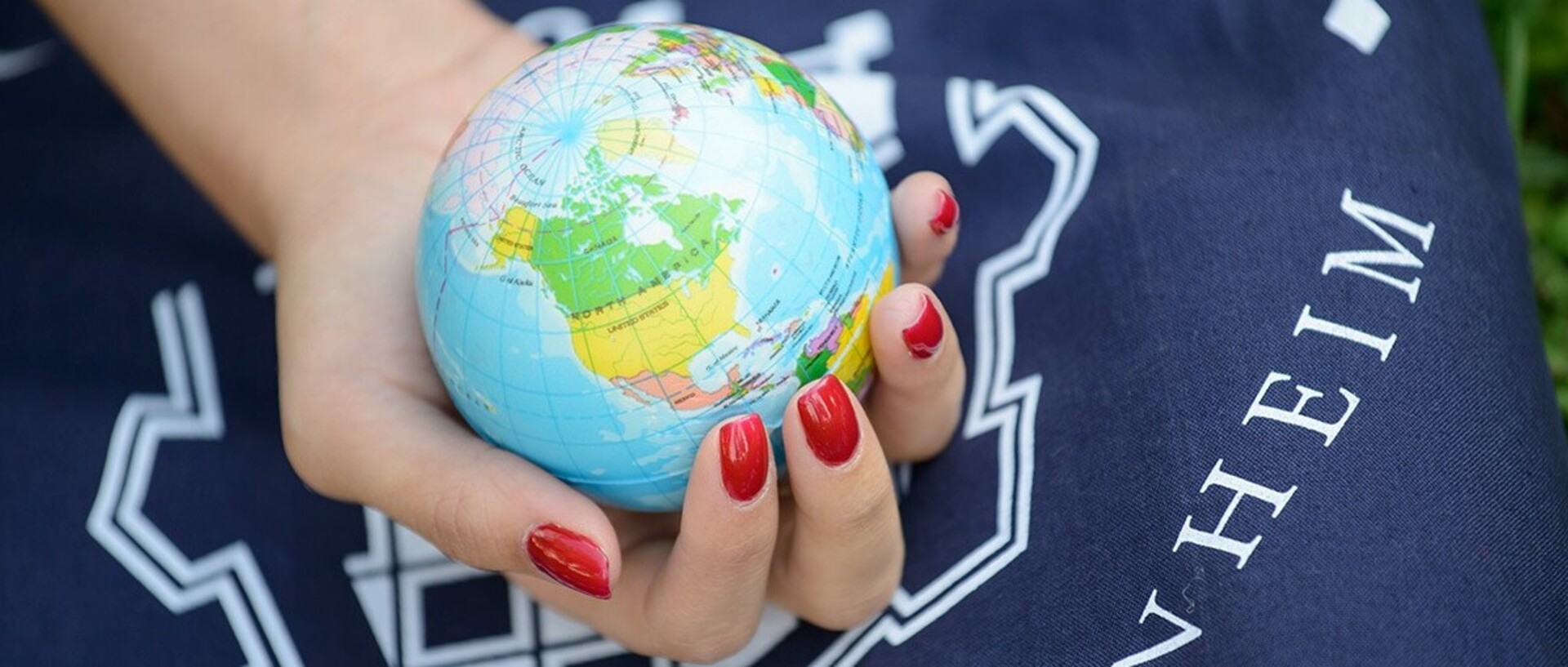 A person with red-painted fingernails holds a small globe in his hand. Underneath is a blue bag with the logo of the University of Mannheim printed on it.