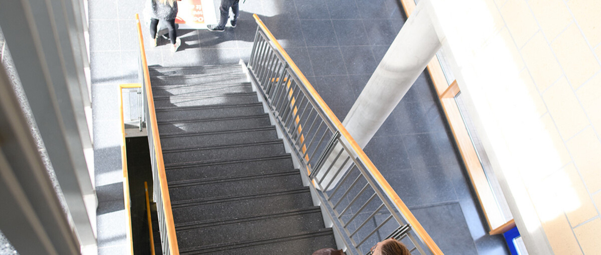 Aufnahme einer Treppe in dem Gebäude B6. Ein Blick von oben in einem modernen Gebäude. Zwei Personen steigen die Treppe hinauf, während im Hintergrund ein hell beleuchteter Eingangsbereich zu sehen ist.