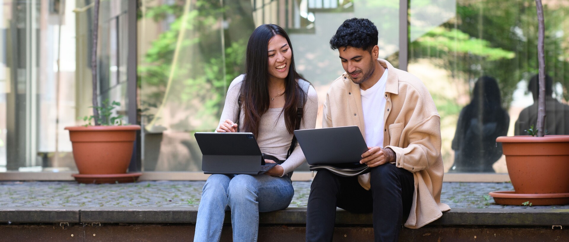 Eine Studentin und ein Student sitzen lachend auf einer Treppe und arbeiten an ihren Laptops.