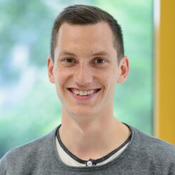 Portrait von Konstantin, einem Studenten der Volkswirtschaftslehre. Er hat kurze braune Haare und trägt ein weißes Oberteil mit schwarzem Saum, darüber trägt er einen hellgrauen Pullover. Konstantin lächelt in die Kamera.