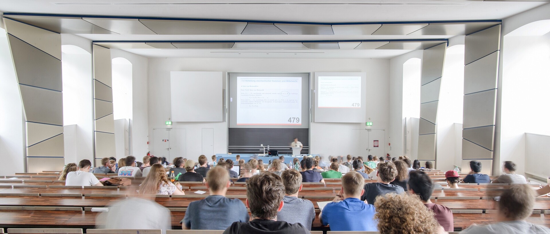 View from the back of lecture hall SO108 in the east wing of the palace. Many students are sitting in the lecture hall and looking at the screen at the front of the lecture hall. The room features design elements in various shades of grey and dark brown tables.