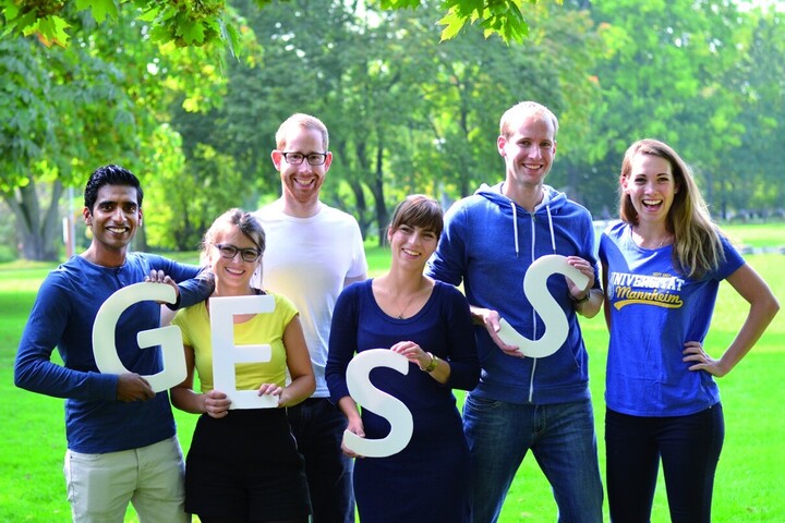 In einem Park stehen sechs Personen die die Buschstaben „G“, „E“ und zweimal „S“ halten. Alle lächeln in die Kamera. 