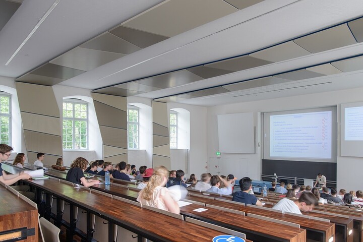 Aufnahme aus dem Hörsaal SO 108 im Ostflügel der Universität Mannheim. In dem Hörsaal sitzen viele Studierende. Die Wände des Hörsaals sind weiß mit grauen Elementen, die Tische sind dunkelbraun.