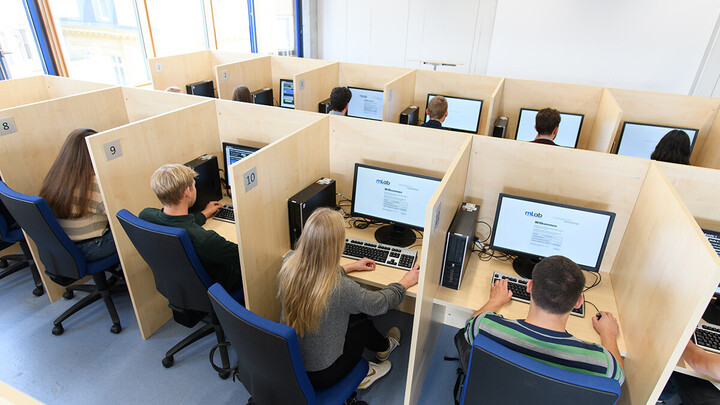 Der helle Raum des mLabs mit mehreren Arbeitskabinen, in denen Personen an Computern arbeiten. Große Fenster im Hintergrund lassen viel Tageslicht herein.