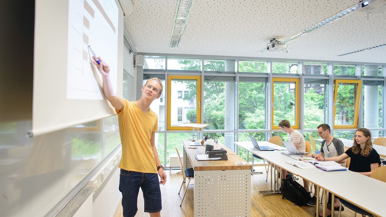 Einblick in einen Übungsraum. Am Whiteboard steht ein Student und erklärt etwas, dabei deutet er auf das Whiteboard. Ihm gegenüber sitzen drei Studierende an Tischen. Auf den Tischen vor einem Studenten steht ein Laptop und zwei Ordner liegen geöffnet vor den anderen beiden.