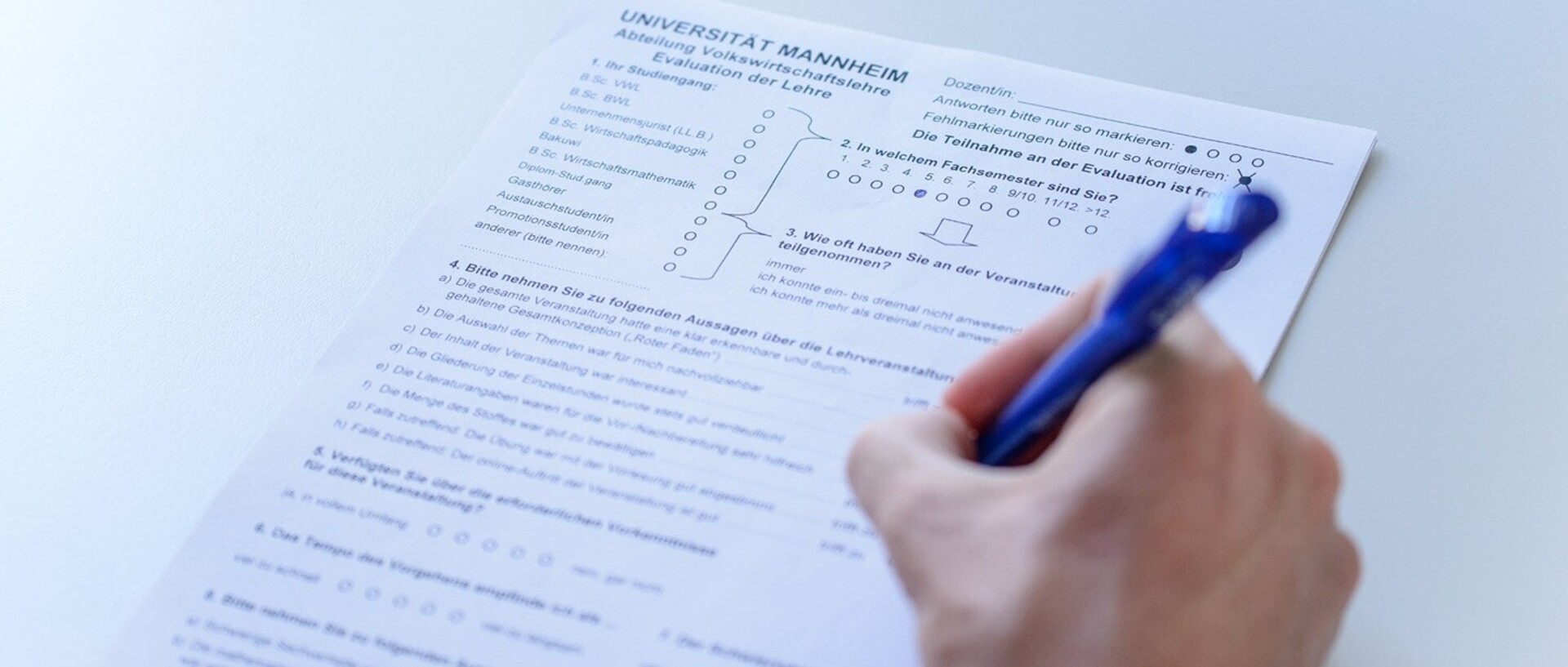 Ein Fragebogen zur Lehrevaluation wird mit einem blauen Stift ausgefüllt. Die Hand der Person, welche den Bogen ausfüllt, befindet sich am rechten Bildrand.