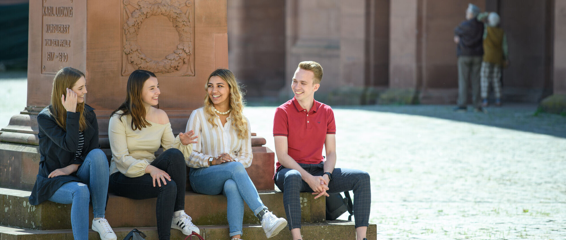 Drei Studentinnen und ein Student sitzen am Fuß einer der Skulpturen auf dem Ehrenhof und unterhalten sich.