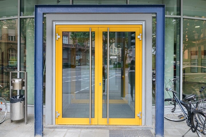 View of the entrance door to the Department of Economics building. The frame of the door is yellow, the door itself and the facade are made of glass. There is a dark blue frame around the door as a decorative element. On the right-hand side of the picture is a black bicycle.
