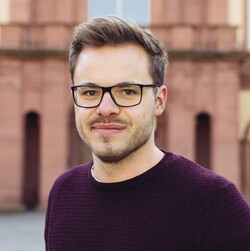 Portrait von Paul Mannschreck, einem Masterstudenten der VWL. Er hat kurze braune Haare, trägt eine Brille und einen dunkelroten Pullover. 