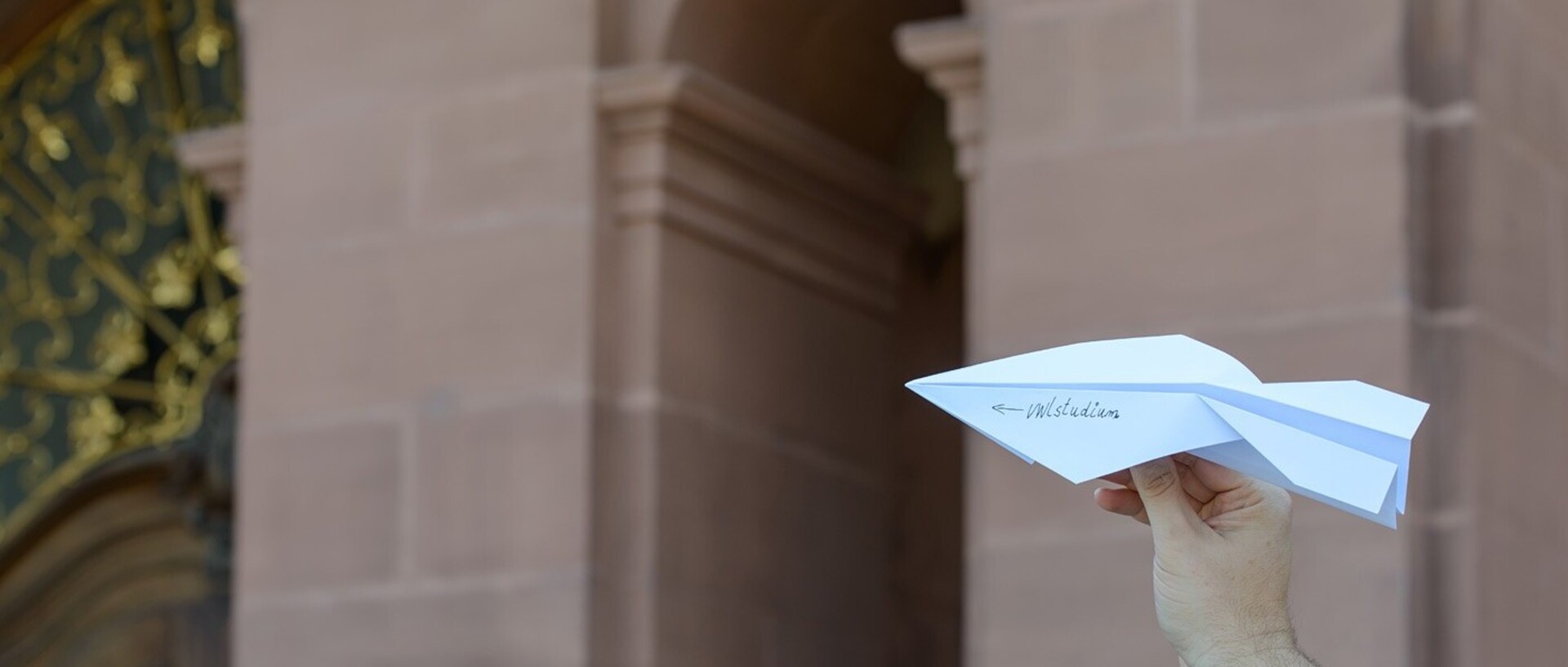 Papierflieger mit der Aufschrift „VWL Studium“ wird vor das Schloss gehalten.