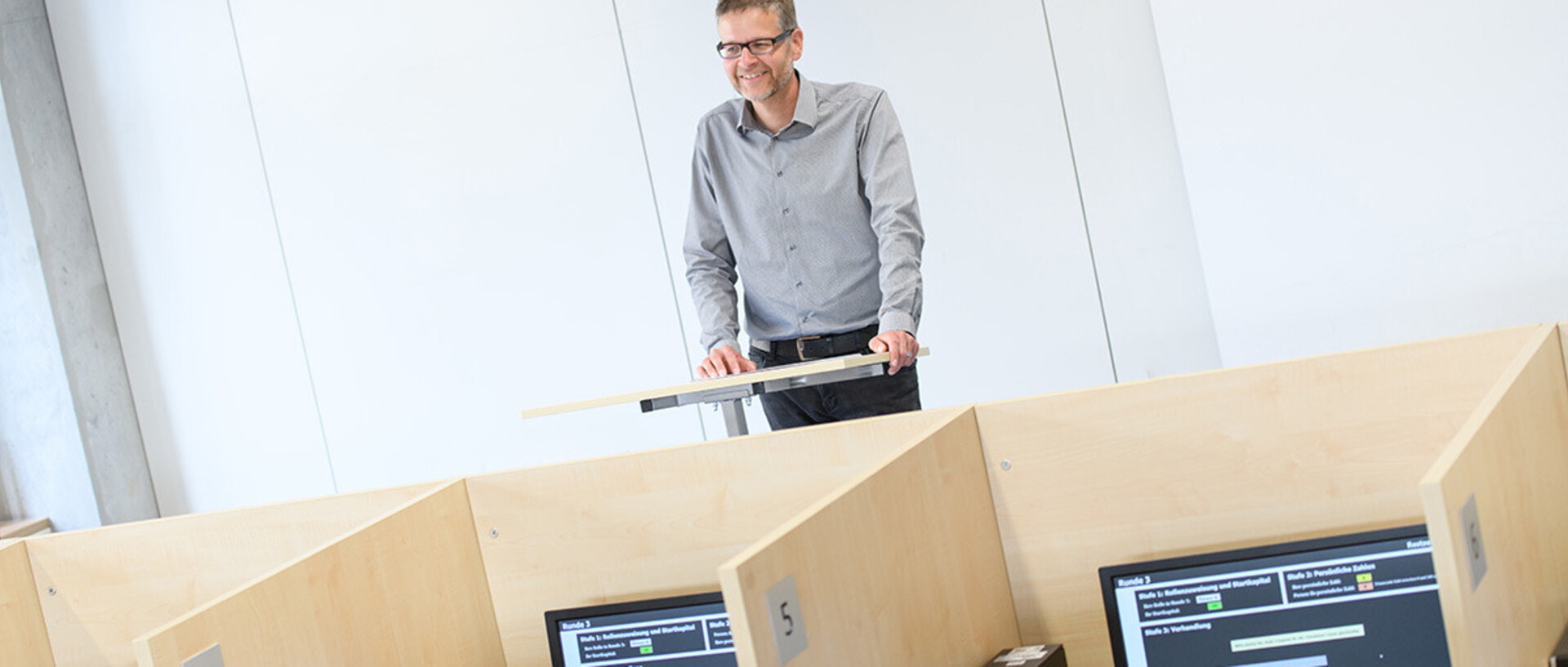 Professor Orzen steht lächelnd vor der Reihe von Computerarbeitsplätzen in einem Raum mit abgetrennten Kabinen. Der Raum vermittelt eine strukturierte und konzentrierte Arbeitsumgebung.