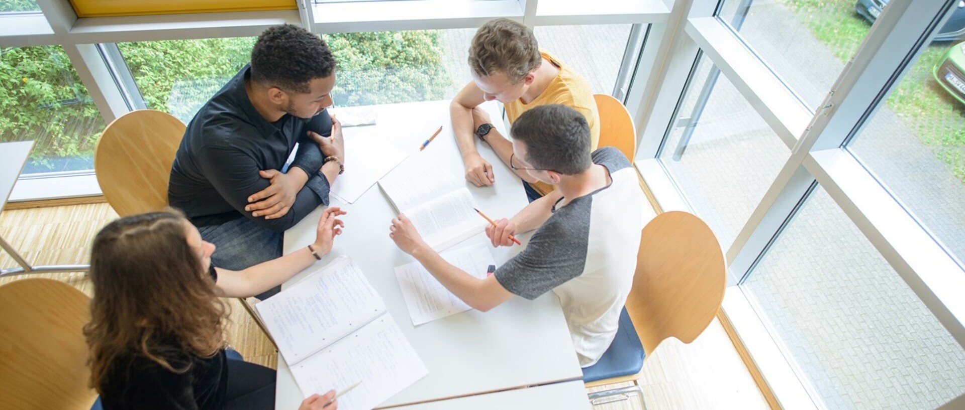 A female student and three male students are sitting around a table in a lecture room. They are bending over notes.