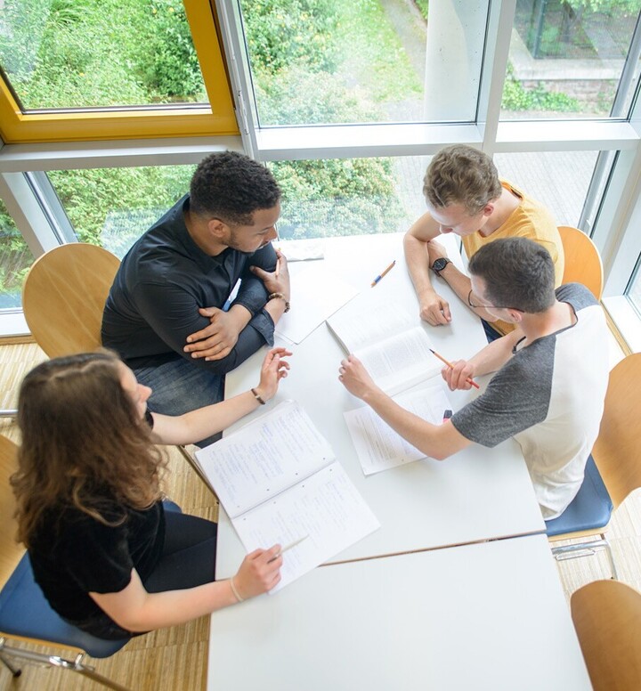 Eine Studentin und drei Studenten sitzen in einem Übungsraum um einen Tisch. Sie arbeiten an ihren Mitschriften.