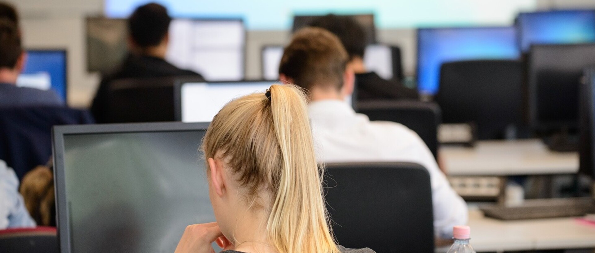 VWL PC-Pool. Einige Studierende sitzen vor den Bildschirmen, im Zentrum des Bildes sitzt eine blonde Frau mit Zopf und schaut auf einen Bildschirm.