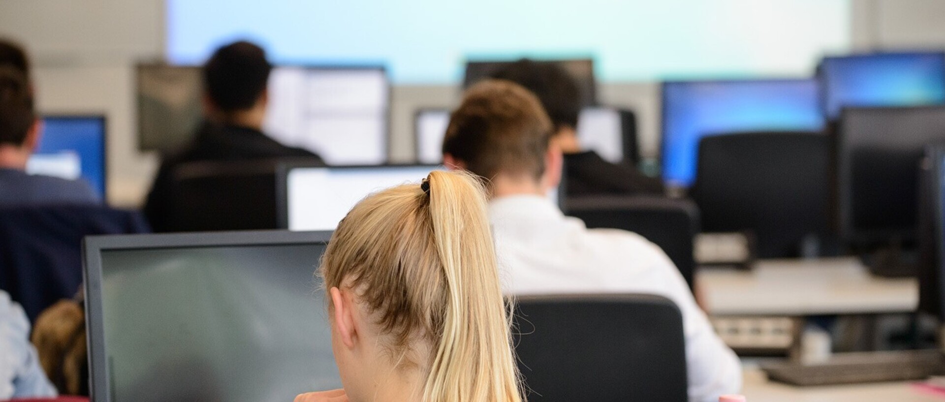 VWL PC pool. Some students are sitting in front of the screens, in the centre of the picture is a blonde woman with a pigtail looking at a screen.