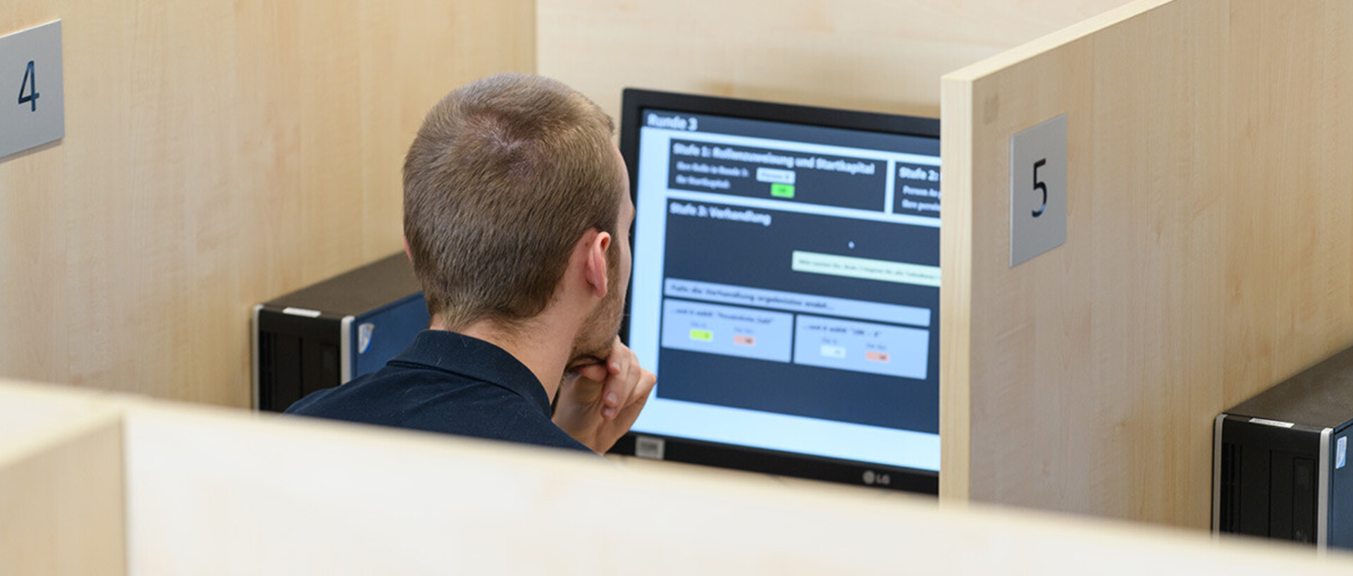 Ein Teilnehmer sitzt in einer abgetrennten Arbeitskabine mit der Nummer 4 und blickt konzentriert auf einen Computerbildschirm. Der Bildschirm zeigt eine Benutzeroberfläche mit Auswahloptionen und Textfeldern.