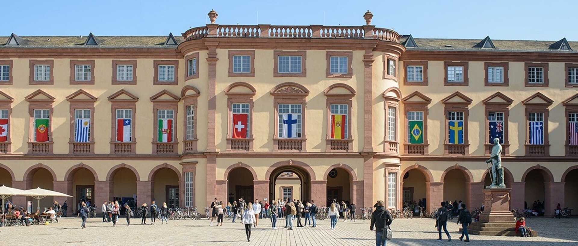 East wing of the University of Mannheim with many flags of different countries on the windows on the first floor. Many people walk across the „Ehrenhof“.