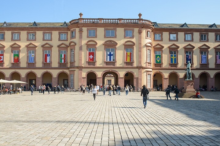 East wing of the University of Mannheim with many flags of different countries on the windows on the first floor. Many people walk across the “Ehrenhof”