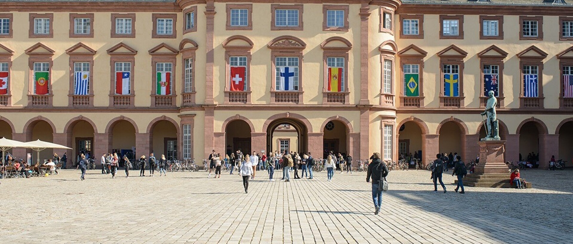 East wing of the University of Mannheim with many flags of different countries on the windows on the first floor. Many people walk across the „Ehrenhof“.