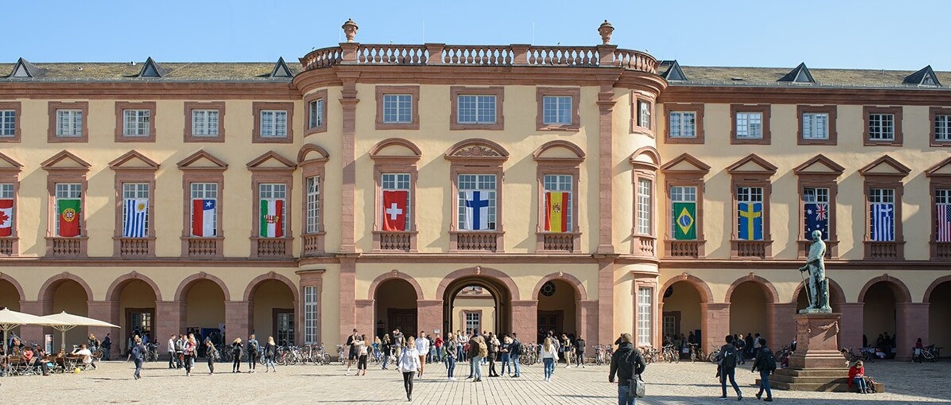 Ostflügel der Universität Mannheim mit vielen Flaggen verschiedener Länder an den Fenstern im ersten Stock. Über den Ehrenhof laufen viele Menschen.