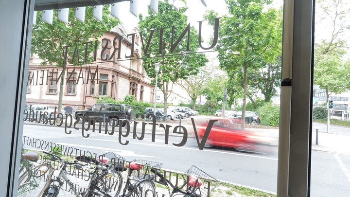 View through a pane of the Economics department building. The glass pane reads: University of Mannheim; Disposition Building; Faculty of Law and Economics; Economics Department Building.