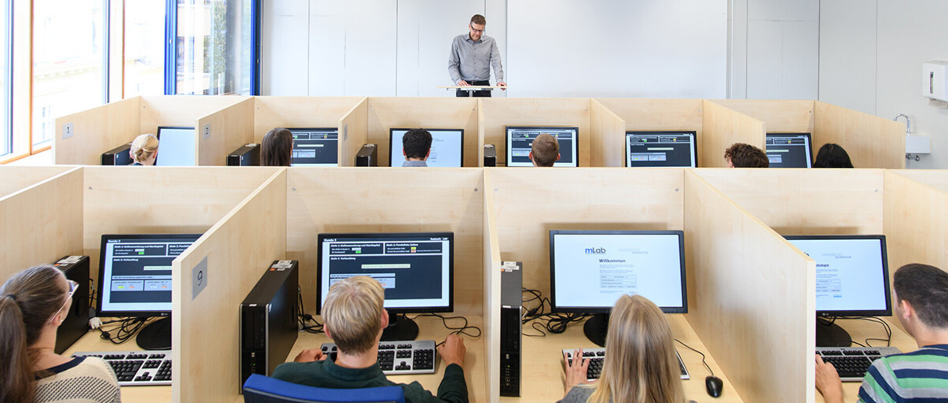 Aufnahme aus dem mLab. Studierende sitzen hinter Bildschirmen, die Arbeitsplätze sind durch Holzwände voneinander abgetrennt. Am Pult vorne steht Professor Orzen. 