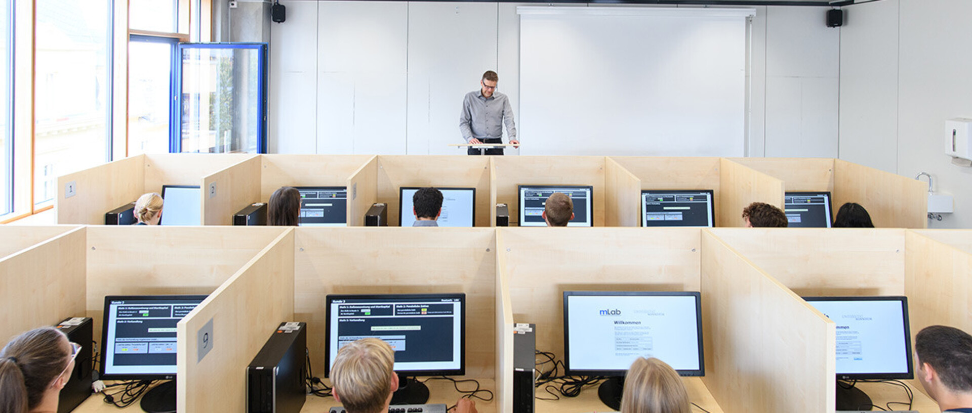 Aufnahme aus dem mLab. Studierende sitzen hinter Bildschirmen, die Arbeitsplätze sind durch Holzwände voneinander abgetrennt. Am Pult vorne steht Professor Orzen.