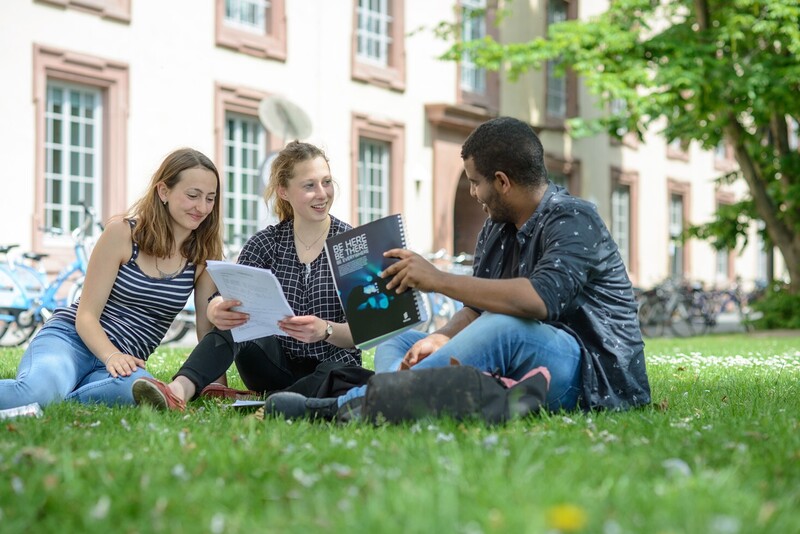 Zwei Studentinnen und ein Student sitzen auf einer Wiese vor dem Mannheimer Schloss. Sie tragen kurzärmlige Oberteile und halten Papierblöcke in ihren Händen.