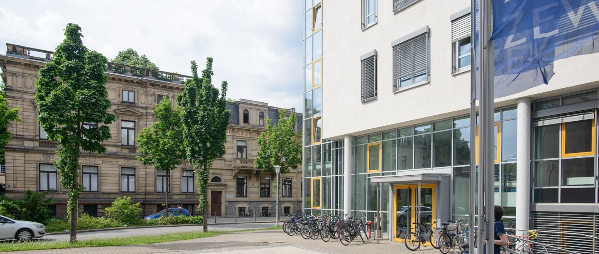 Blick auf das Abteilungsgebäude der Abteilung Volkswirtschaftslehre. Auf der linken Bildseite stehen Bäume vor anderen Gebäuden der Universität Mannheim im Block L5. Rechtsseitig befindet sich das VWL-Gebäude, vor dem einige Fahrräder stehen. In der rechten oberen Ecke wehen die dunkelblauen Fahnen des ZEW.