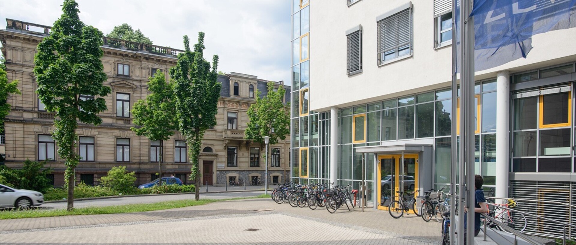 View of the Department of Economics building. On the left side of the picture there are trees in front of another university building. On the right side is the economics building in front of which some bicycles are parked. In the upper right corner, the dark blue flags of the ZEW are flying.