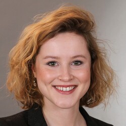 Portrait von Elisabeth Kurz, einer Masterstudentin der VWL. Sie hat kinnlange blonde Locken und trägt ein rotes Oberteil und darüber einen schwarzen Blazer.