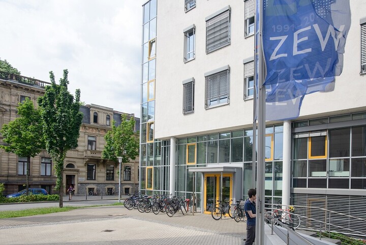 Blick auf das Gebäude der Abteilung Volkswirtschaftslehre. Auf der linken Bildseite stehen Bäume vor anderen Gebäuden der Universität im Block L5. Rechtsseitig befindet sich das VWL-Gebäude, vor dem einige Fahrräder stehen. In der rechten oberen Ecke wehen die dunkelblauen Fahnen des ZEW.