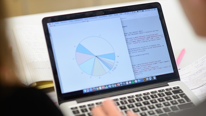 View of an opened laptop. On the left side of the screen is a pie chart, on the right side and in the background is the code for the chart. Two people are looking at the laptop.